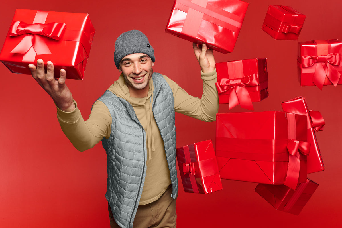 A smiling man wearing a gray beanie, tan hoodie, and light blue quilted vest holds two red gift boxes with ribbons against a solid red background, with more red gift boxes floating around him. The North Face® Circular Rib Beanie, Embroidered