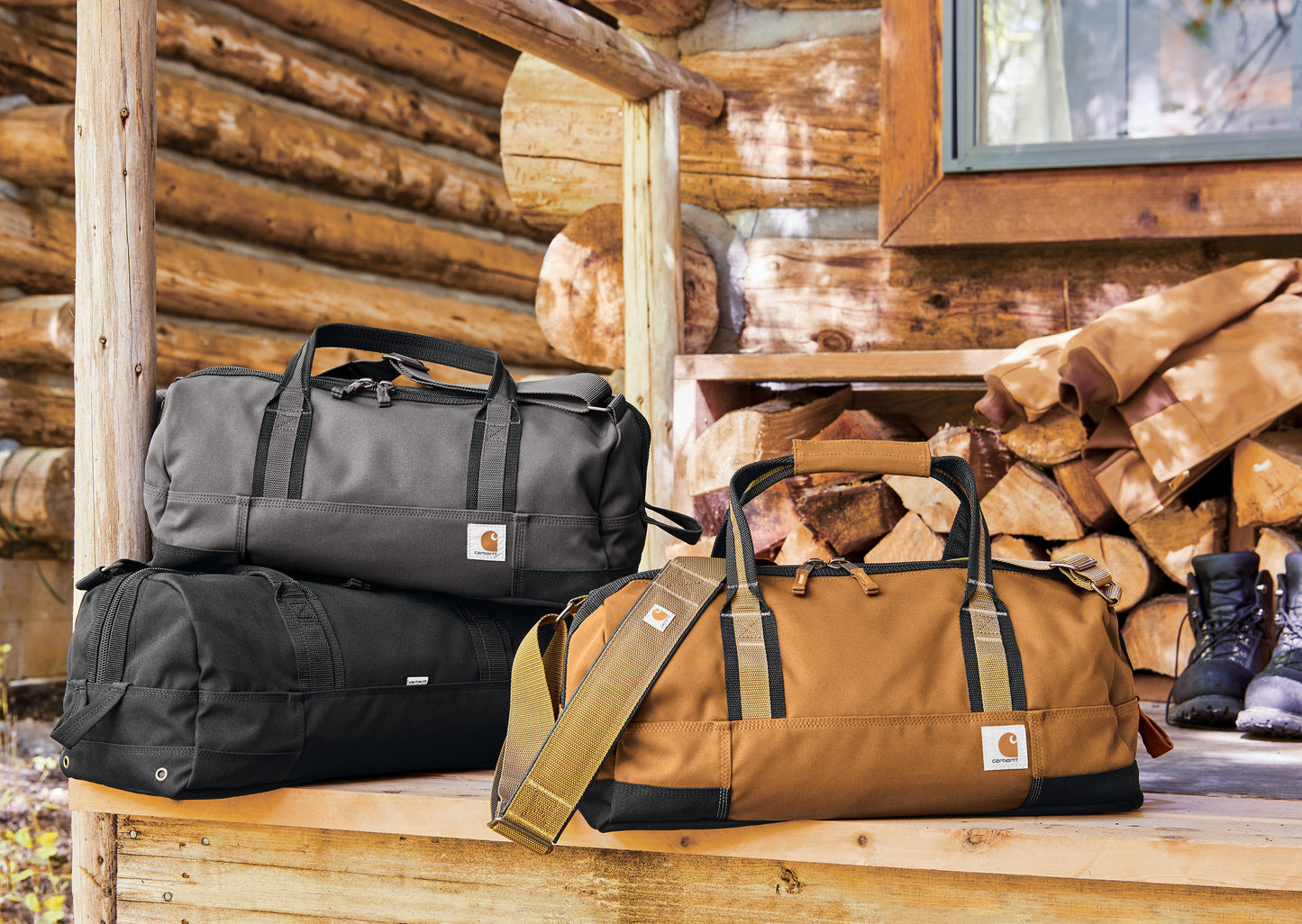 Three bags (black, brown, and black) on a wooden surface with a log cabin background. embroidered Carhartt® Foundry Series 20” Duffel