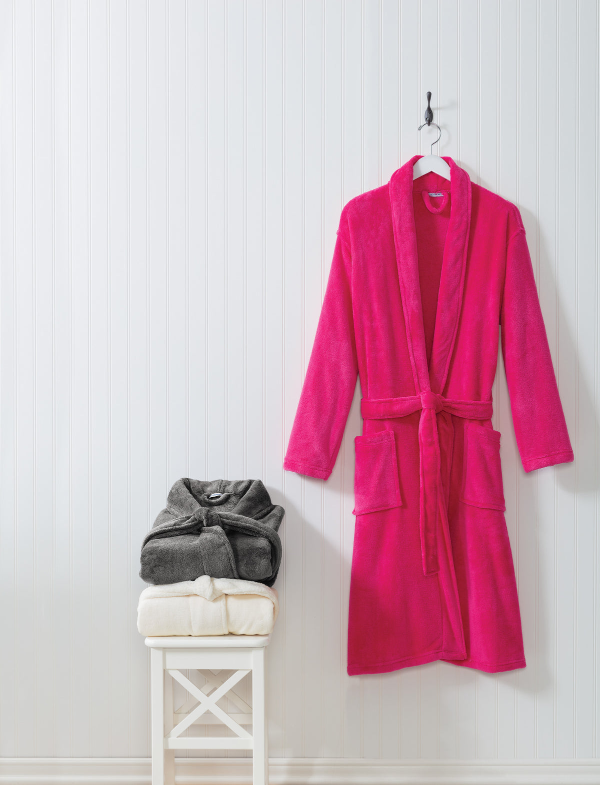 A bright pink plush bathrobe hanging on a white hanger against a white paneled wall, with folded gray and cream towels stacked on a small white stool beside it. Port Authority® Plush Microfleece Shawl Collar Robe, Embroidered