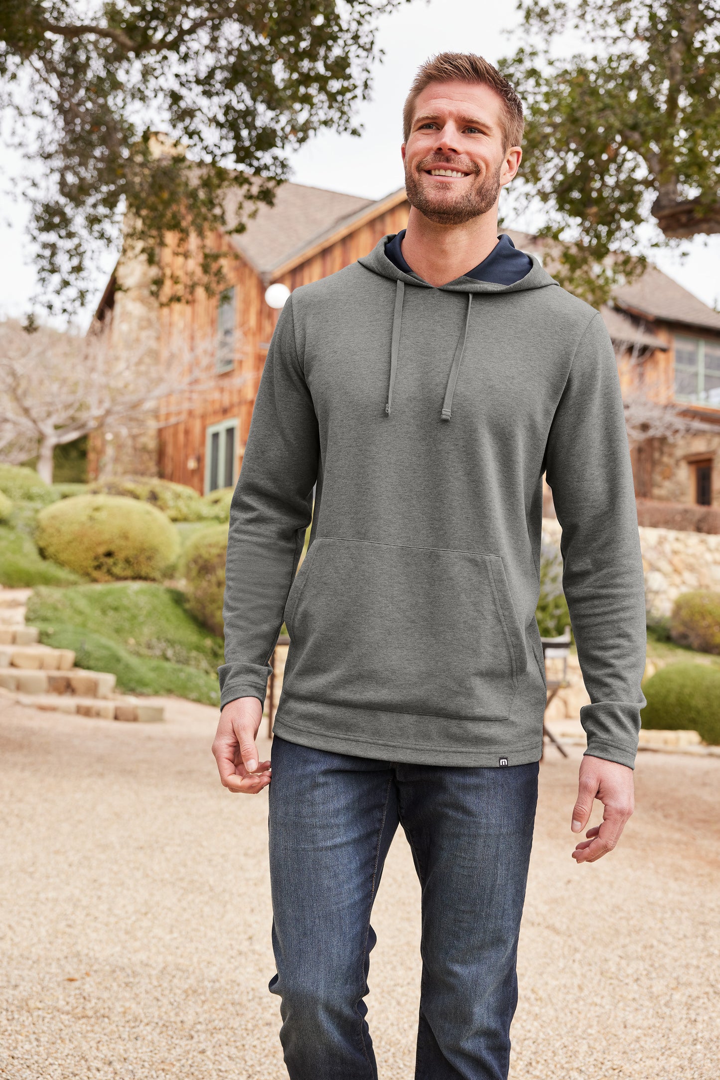 Man wearing a gray hoodie and blue jeans standing outdoors with a house in the background TravisMathew Coveside Hoodie, Embroidered