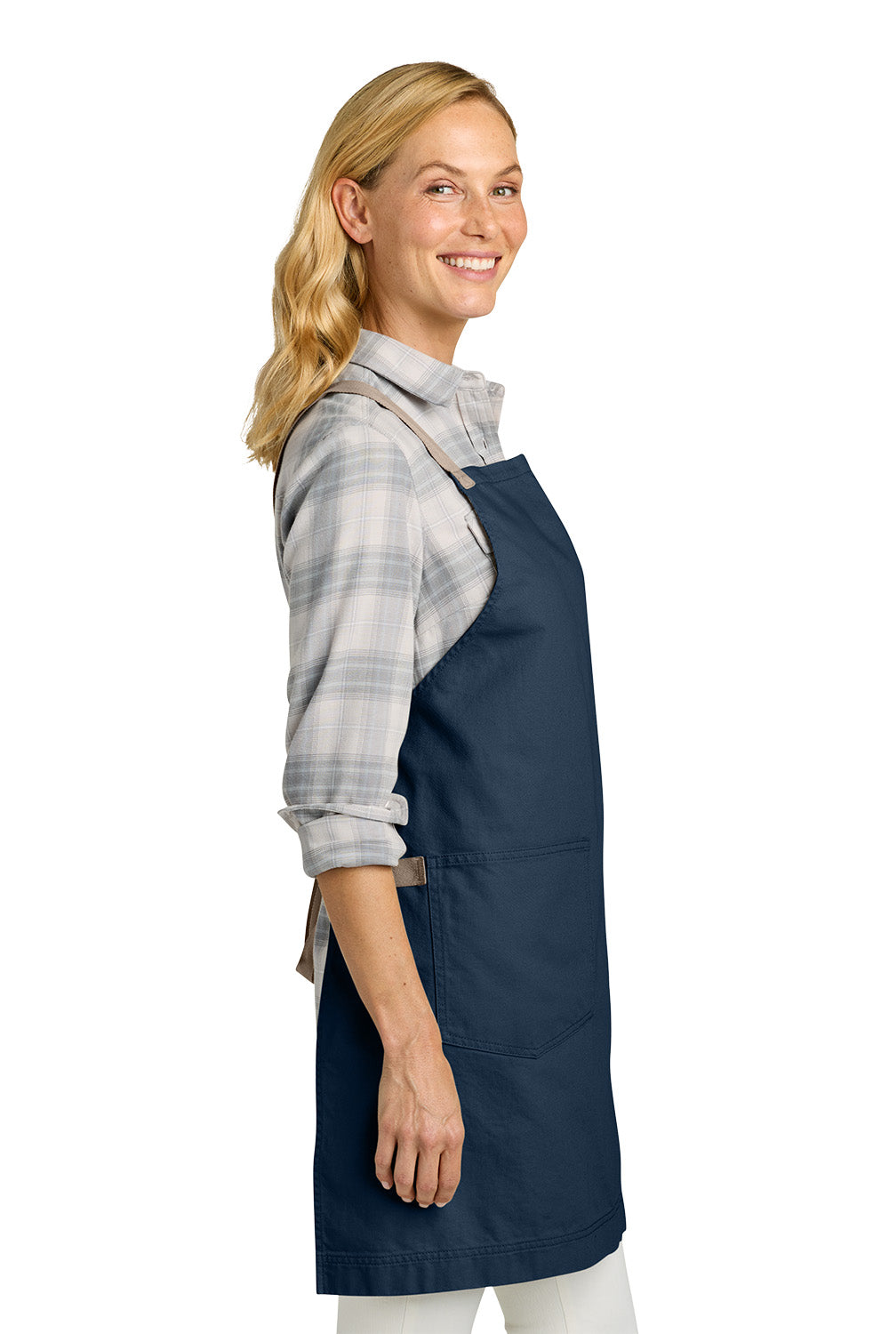 A woman wearing a navy blue bib apron over a gray and white plaid flannel shirt, shown in profile against a solid white background. Port Authority® Signature Heavy Twill Crossback Apron, Embroidered