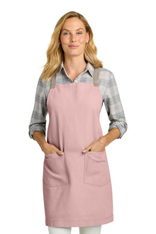 A woman modeling a dusty pink bib apron with two front pockets over a gray plaid flannel shirt, photographed against a solid white background. Port Authority® Signature Heavy Twill Crossback Apron, Embroidered