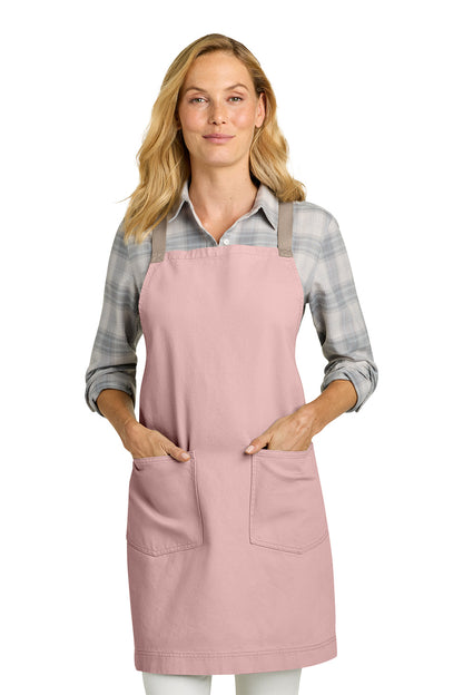 A woman modeling a dusty pink bib apron with two front pockets over a gray plaid flannel shirt, photographed against a solid white background. Port Authority® Signature Heavy Twill Crossback Apron, Embroidered