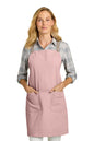 A woman modeling a dusty pink bib apron with two front pockets over a gray plaid flannel shirt, photographed against a solid white background. Port Authority® Signature Heavy Twill Crossback Apron, Embroidered