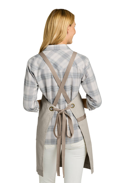 Woman wearing a pink apron with a brown strap over a plaid shirt on a white background. Port Authority® Signature Heavy Twill Crossback Apron, Embroidered