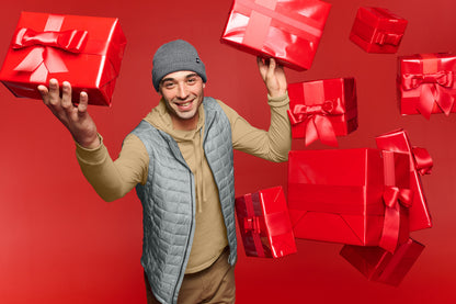 A smiling man wearing a gray beanie, tan hoodie, and light blue quilted vest holds two red gift boxes with ribbons against a solid red background, with more red gift boxes floating around him. The North Face® Circular Rib Beanie, Embroidered