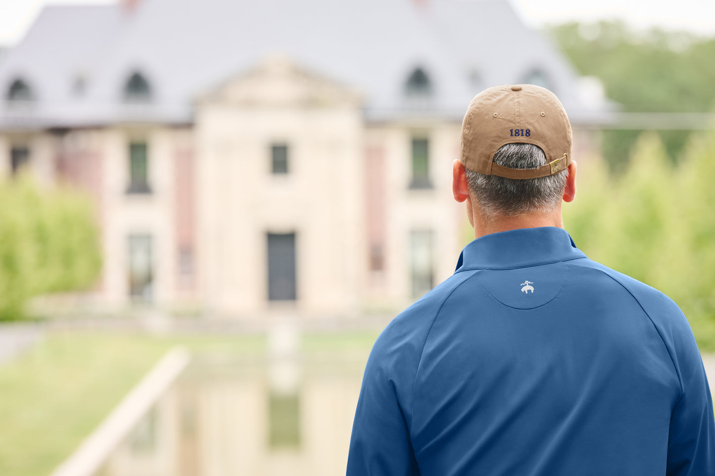 Man wearing a blue shirt and tan cap standing in front of a large house. Brooks Brothers® Luxe Performance 1/4-Zip, Embroidered