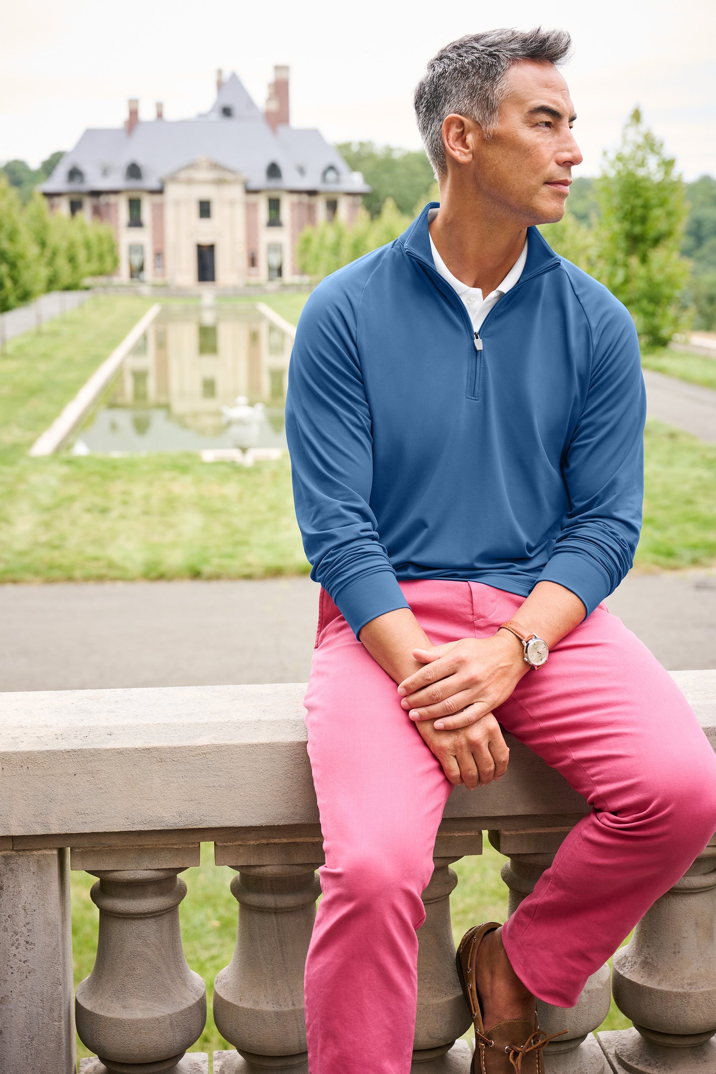 Man sitting on a stone bench wearing a blue sweater and pink pants with a scenic background. Brooks Brothers® Luxe Performance 1/4-Zip, Embroidered