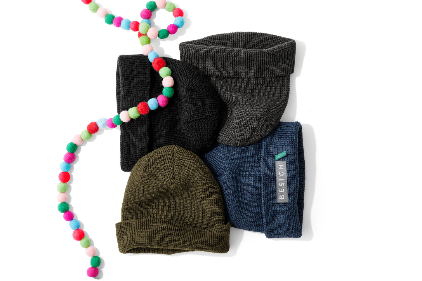 Four ribbed knit beanie hats in black, charcoal gray, olive green, and navy blue, arranged on a white background with a colorful pom-pom bead garland draped over them. A small white tag on the navy beanie reads REACH. Port Authority® Thermal Knit Cuff Beanie, Embroidered