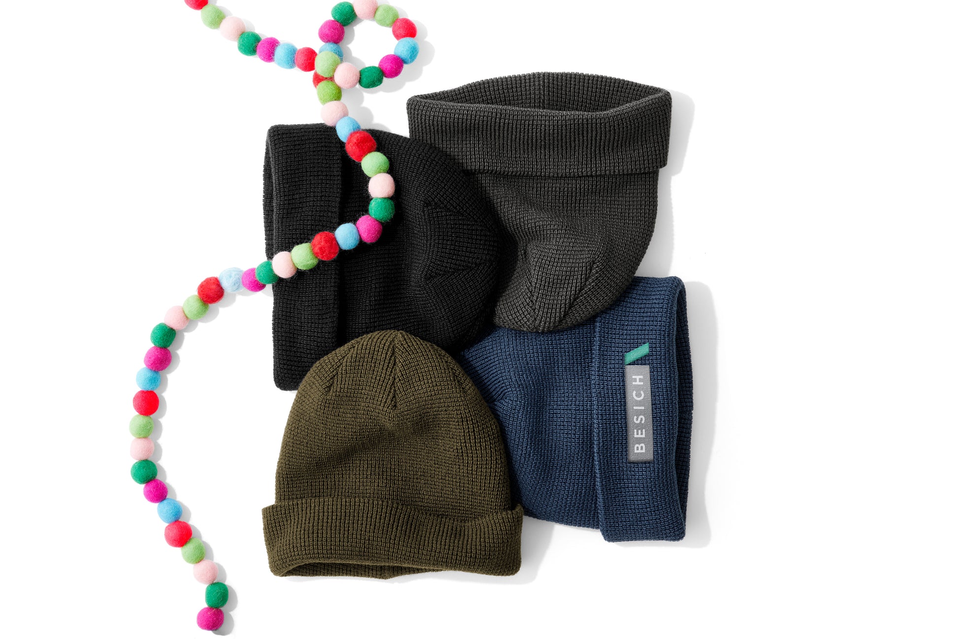 Four ribbed knit beanie hats in black, charcoal gray, olive green, and navy blue, arranged on a white background with a colorful pom-pom bead garland draped over them. A small white tag on the navy beanie reads REACH. Port Authority® Thermal Knit Cuff Beanie, Embroidered