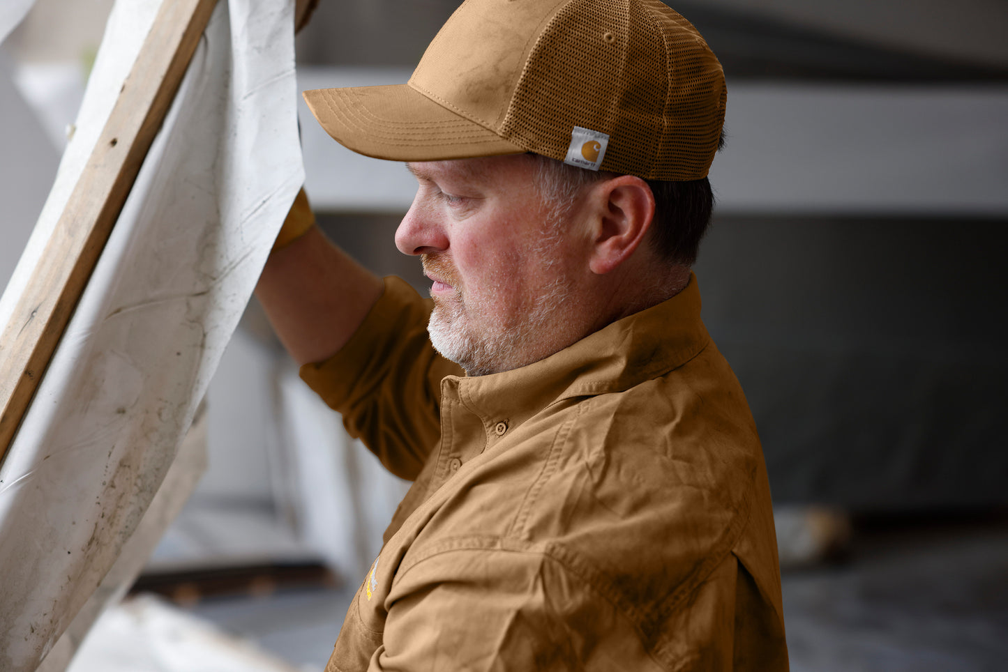 Man wearing a brown cap and jacket, working with fabric or paper. Carhartt® Flexfit 110® Mesh Back Cap, Embroidered