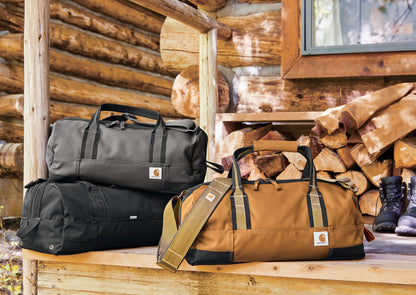 Three bags (black, brown, and black) on a wooden surface with a log cabin background. embroidered Carhartt® Foundry Series 20” Duffel