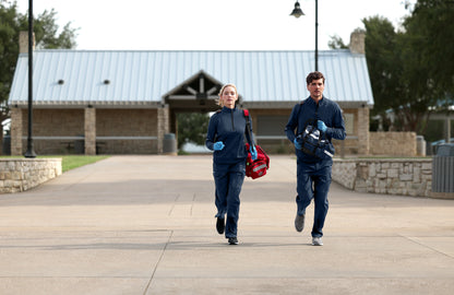Two people walking outdoors on a paved path with a building and trees in the background. Carhartt® FLD & RSCU 1/2-Zip Mid-Layer, Embroidered