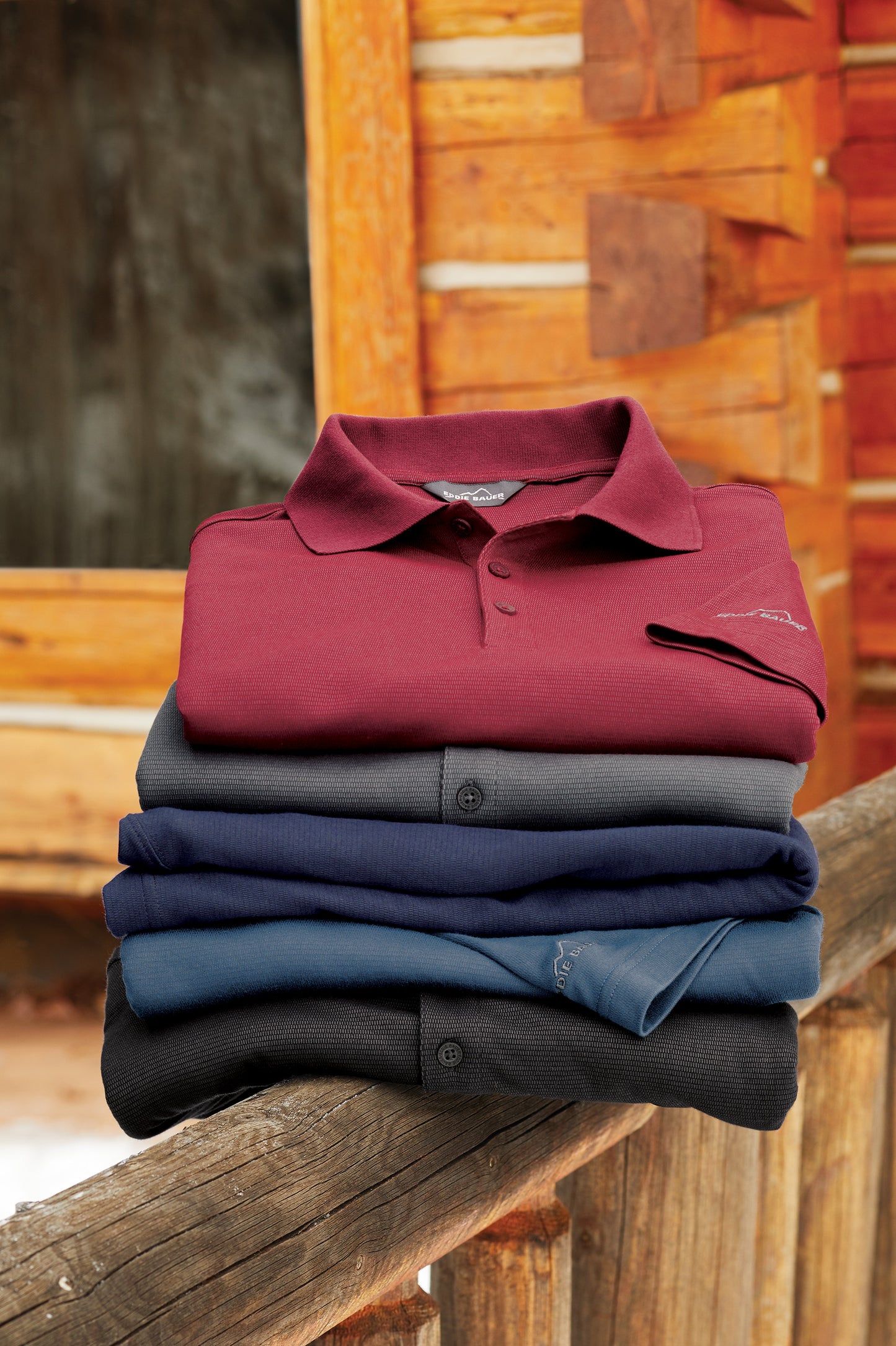 Stack of folded polo shirts in red, blue, and black on a wooden surface with a rustic background. Eddie Bauer® Performance Polo, Embroidered