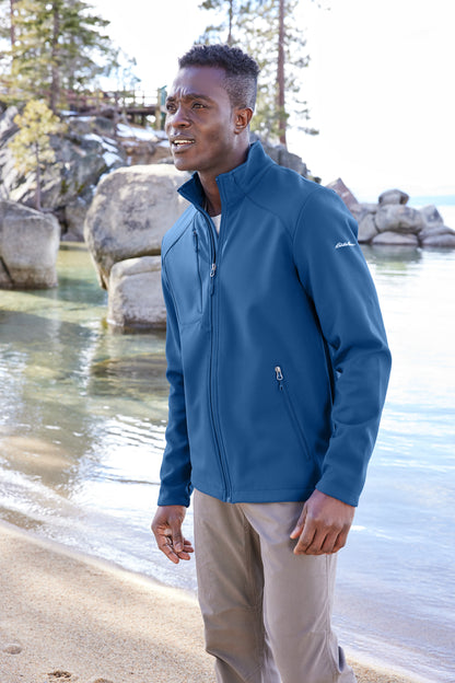 Man wearing a blue jacket standing by a lake with rocks and trees in the background embroidered Eddie Bauer® Stretch Soft Shell Jacket