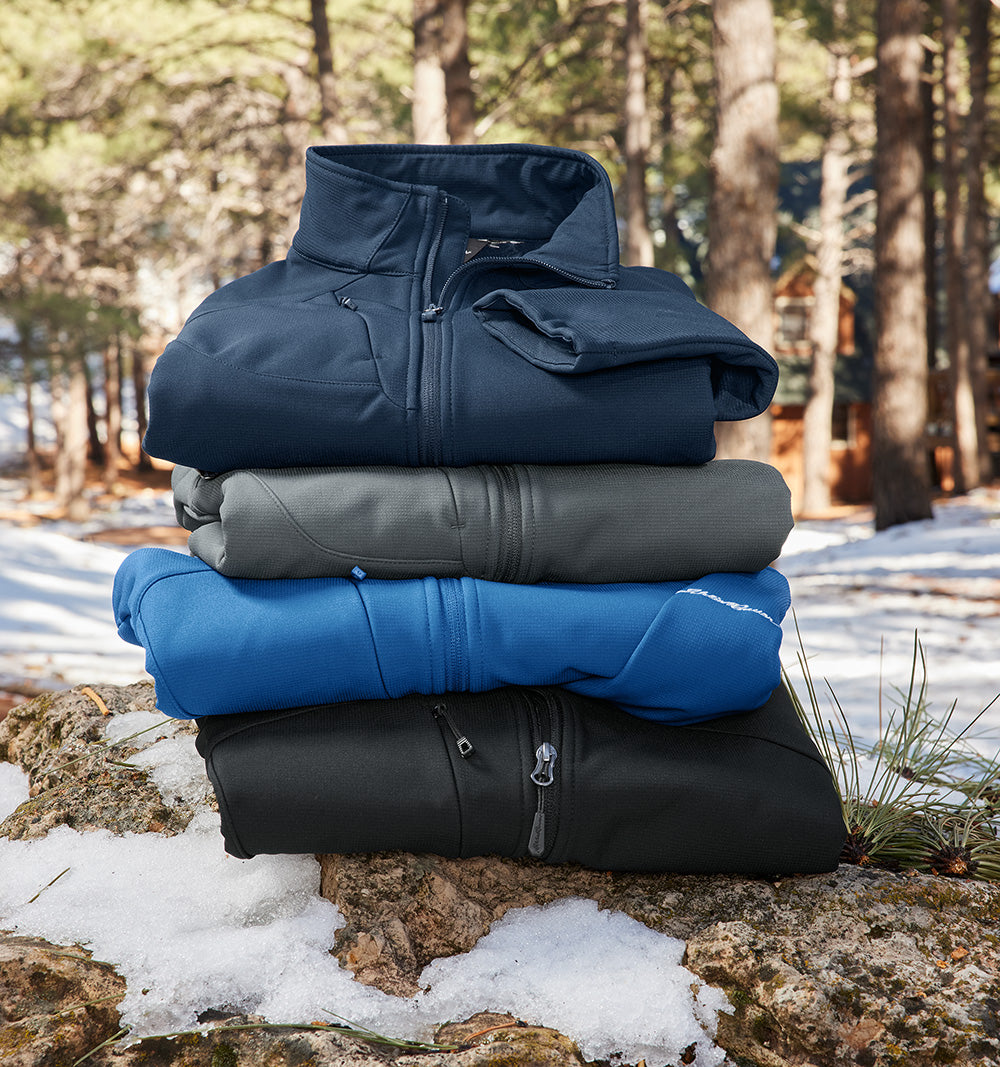 Stack of three insulated jackets in blue, gray, and black on a snowy rock with a forest background. embroidered Eddie Bauer® Stretch Soft Shell Jacket