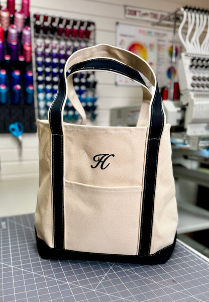 A beige canvas tote bag with black trim and handles, featuring a black embroidered monogram letter 'H' on the front pocket, displayed on a cutting mat inside a sewing or embroidery workshop. Custom Tote, Superior MI Tote, XL, Open Top, Helmsman Stitch Company. 