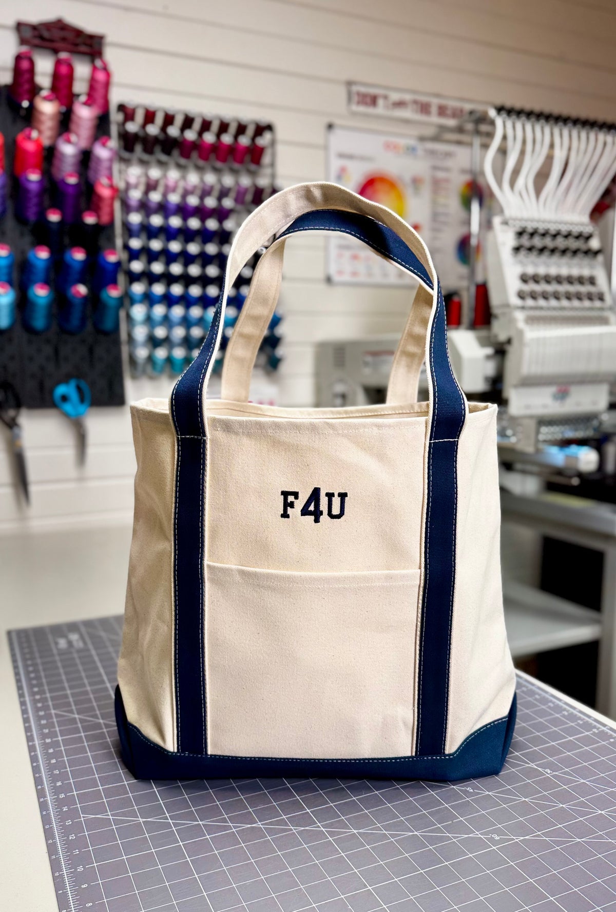 A cream canvas tote bag with navy blue trim and handles, featuring the text 'F4U' embroidered on the front pocket, displayed on a gray cutting mat inside a craft room. Custom Tote, Superior MI Tote, XL, Open Top, Helmsman Stitch Company. 