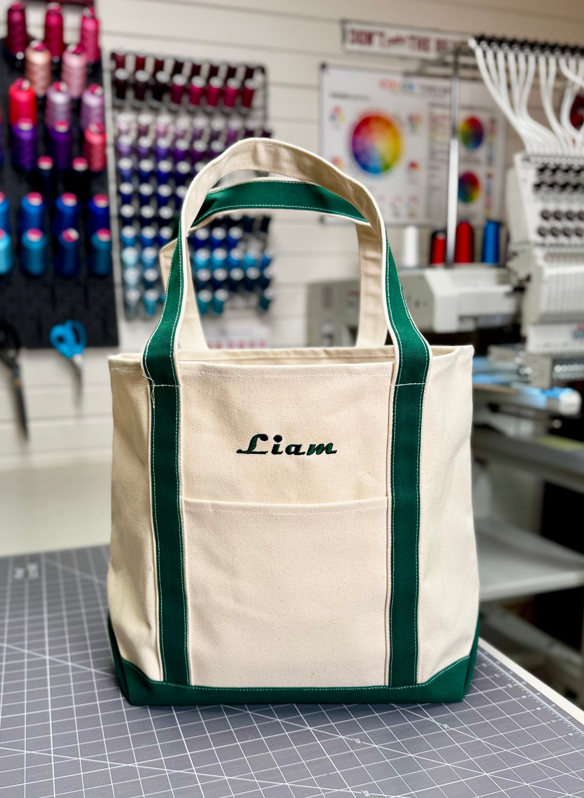 A personalized canvas tote bag with green trim and handles, featuring the name 'Liam' embroidered in black script on the front pocket. The bag is displayed on a cutting mat inside what appears to be an embroidery or sewing workshop. Custom Tote, Superior MI Tote, XL, Open Top, Helmsman Stitch Company. 