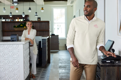 A man in a cream-colored long-sleeve polo shirt with a small pink logo on the left chest stands at a counter in a modern restaurant or cafe, while a woman is visible in the background near the reception desk. Port Authority® Breakwater 1/4-Zip Pullover, Embroidered