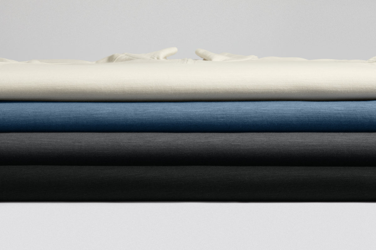 Four folded fabric or upholstery material samples stacked horizontally, showing cream, blue, charcoal gray, and black in a clean studio setup. Port Authority® Breakwater 1/4-Zip Pullover, Embroidered
