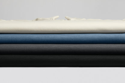 Four folded fabric or upholstery material samples stacked horizontally, showing cream, blue, charcoal gray, and black in a clean studio setup. Port Authority® Breakwater 1/4-Zip Pullover, Embroidered