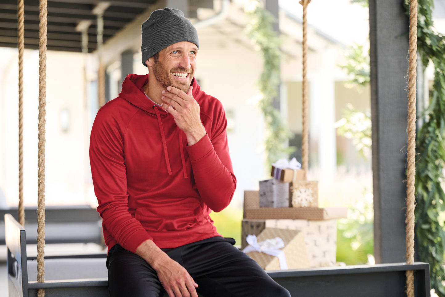Man in a red hoodie and gray beanie sitting on a swing outdoors.New Era® Recycled Cuff Beanie, Embroidered