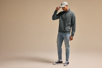 A man modeling a dark gray quarter-zip pullover hoodie, light-wash blue jeans, and a white baseball cap against a solid beige studio background. New Era® Heritage Fleece 1/2-Zip, Embroidered