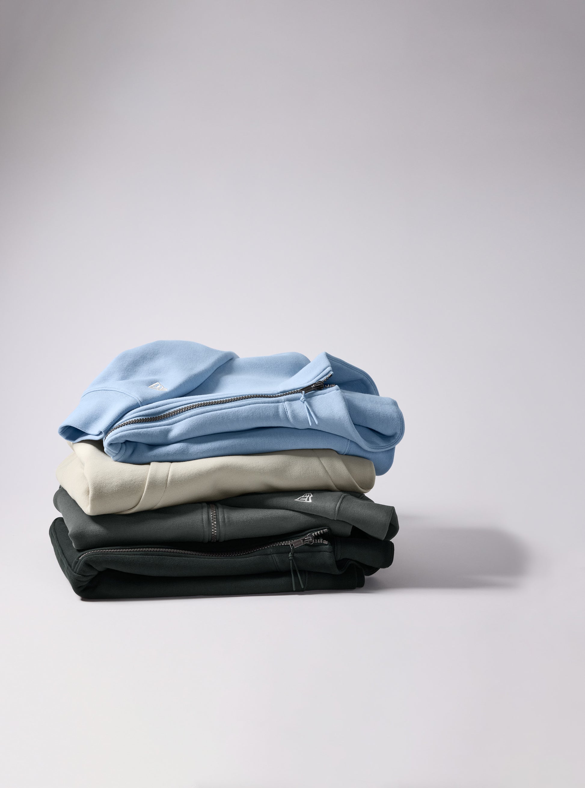 A stack of four folded zip-up sweatshirts in light blue, cream, dark gray, and black, arranged on a light gray background. New Era® Heritage Fleece 1/2-Zip, Embroidered