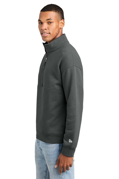 A man wearing a dark gray quarter-zip pullover sweatshirt with a small white New Era logo on the left sleeve cuff, paired with light blue jeans, shown against a plain white background. New Era® Heritage Fleece 1/2-Zip, Embroidered
