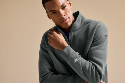 A man wearing a dark gray quarter-zip fleece pullover with a small white embroidered logo on the left sleeve, posed against a solid beige background. New Era® Heritage Fleece 1/2-Zip, Embroidered
