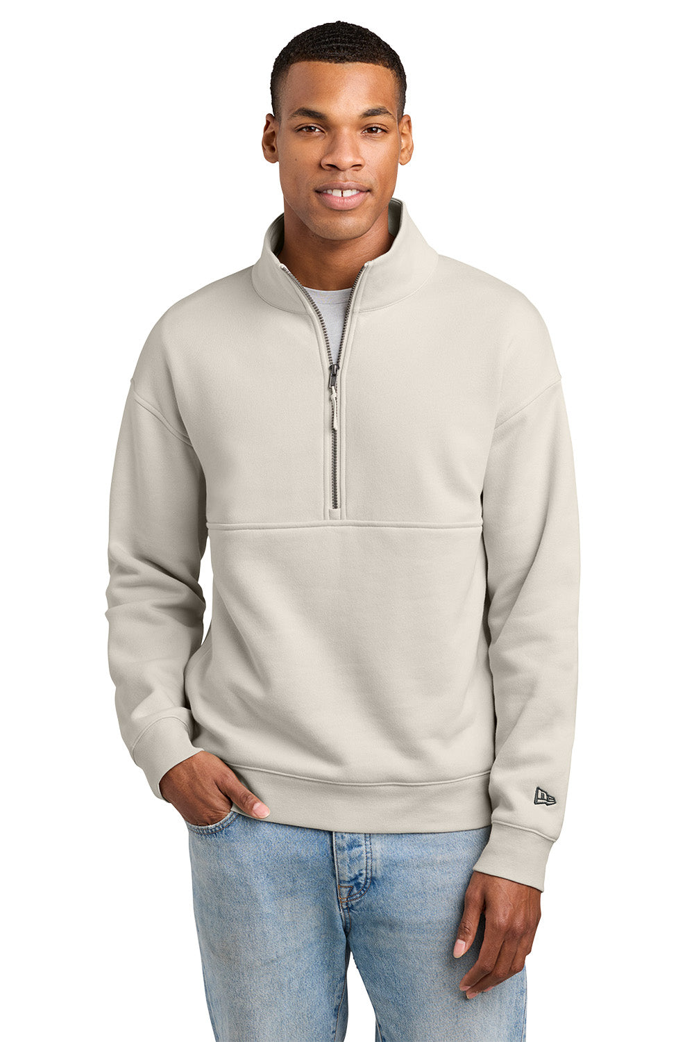 A man wearing a light beige quarter-zip sweatshirt with a mock neck and a horizontal seam across the chest, paired with light blue jeans, against a plain white background.New Era® Heritage Fleece 1/2-Zip, Embroidered