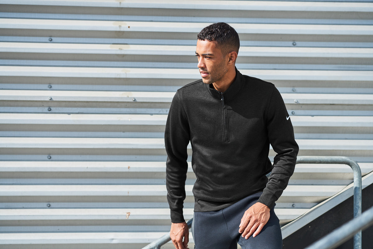 A man wearing a black long-sleeve quarter-zip athletic pullover and dark pants, standing in front of a gray corrugated metal wall. Nike Dri-FIT Corporate 1/2-Zip, Embroidered