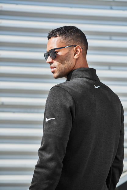 A man in profile wearing a dark charcoal gray Nike pullover and black sunglasses, standing in front of a light gray corrugated metal wall. Nike Dri-FIT Corporate 1/2-Zip, Embroidered