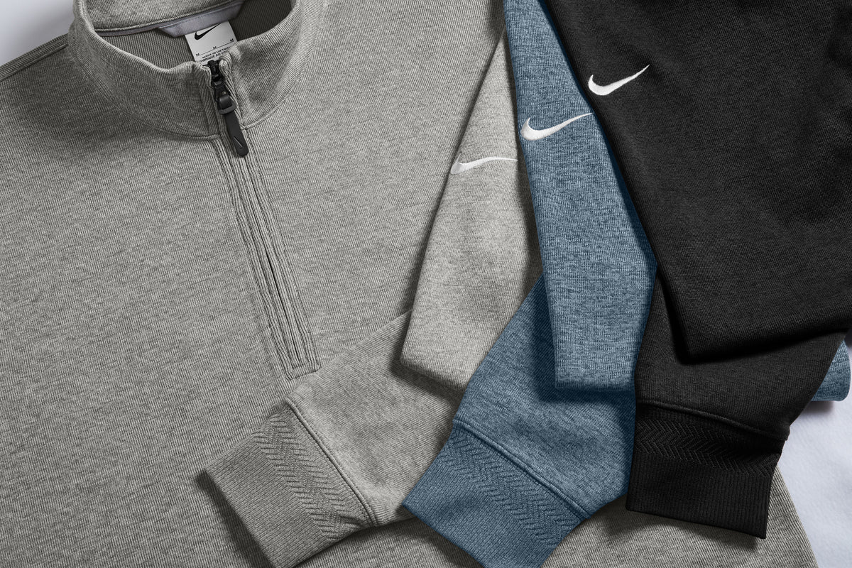 Three Nike quarter-zip pullover sweatshirts arranged diagonally, showing gray, heather blue, and black colorways with white embroidered swoosh logos. Nike Dri-FIT Corporate 1/2-Zip, Embroidered