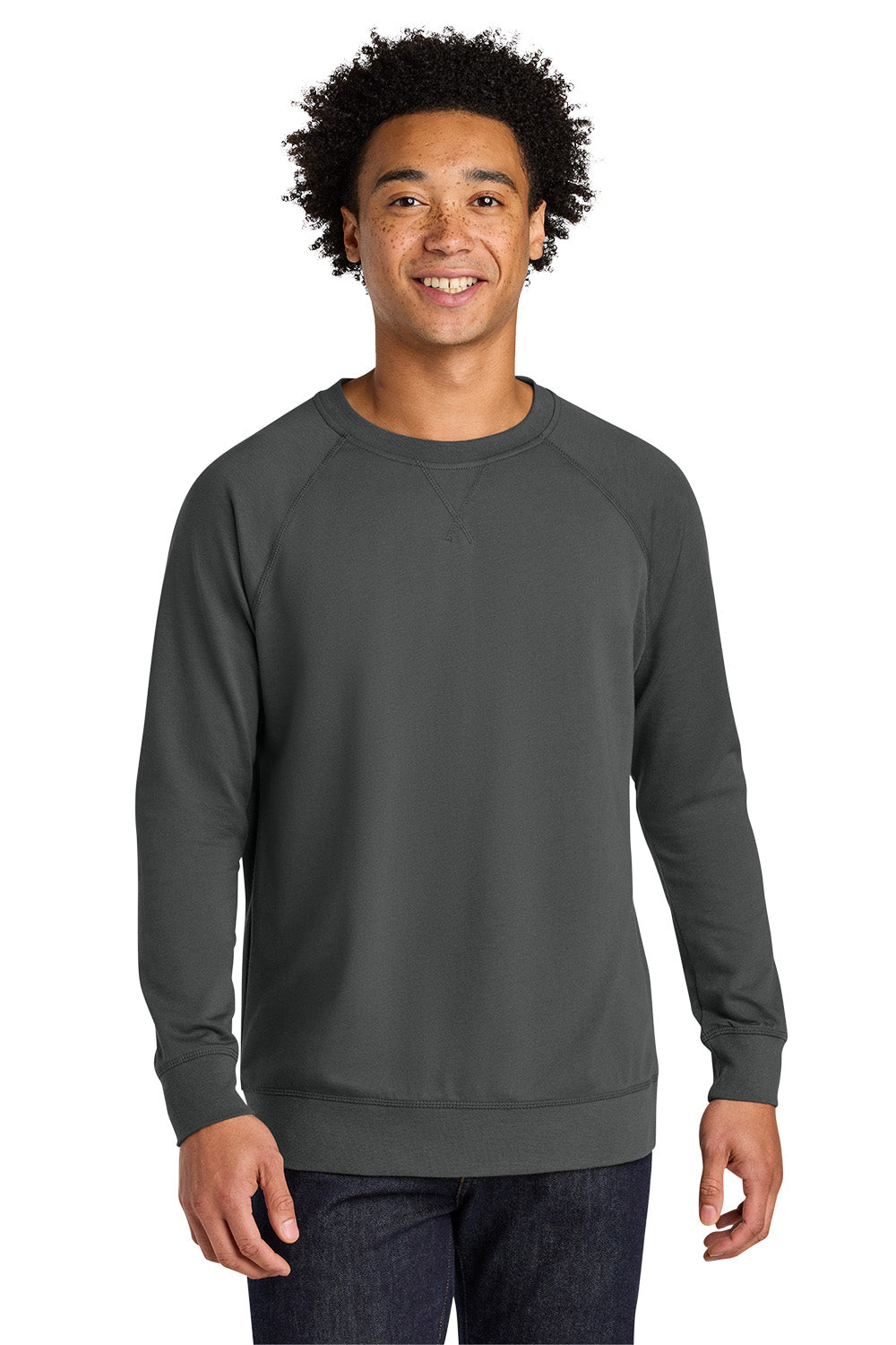 A smiling man with curly black hair wearing a dark gray crewneck sweatshirt and dark blue jeans, standing against a solid white background. Next Level Apparel® Laguna Raglan Sweatshirt, Embroidered