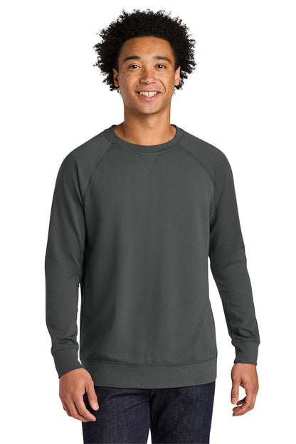 A smiling man with curly black hair wearing a dark gray crewneck sweatshirt and dark blue jeans, standing against a solid white background. Next Level Apparel® Laguna Raglan Sweatshirt, Embroidered