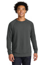 A smiling man with curly black hair wearing a dark gray crewneck sweatshirt and dark blue jeans, standing against a solid white background. Next Level Apparel® Laguna Raglan Sweatshirt, Embroidered