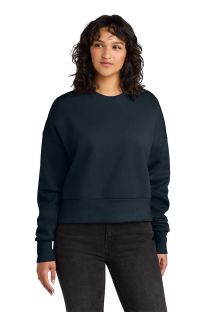 A woman with dark curly hair wearing a black crewneck sweatshirt and black jeans, standing against a solid white background. Next Level Apparel® Women’s Heavyweight Sweatshirt, Embroidered