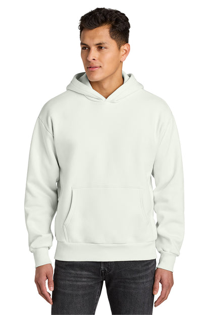A man wearing a light off-white pullover hoodie with a front kangaroo pocket, paired with dark gray jeans, photographed against a solid white background.  Next Level Apparel® Heavyweight Hoodie, Embroidered