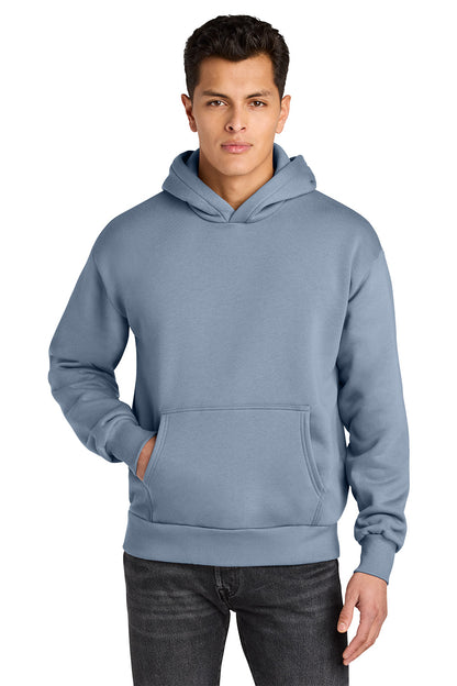 A man wearing a dusty blue pullover hoodie with a front kangaroo pocket, paired with dark gray jeans, photographed against a solid white background. Next Level Apparel® Heavyweight Hoodie, Embroidered