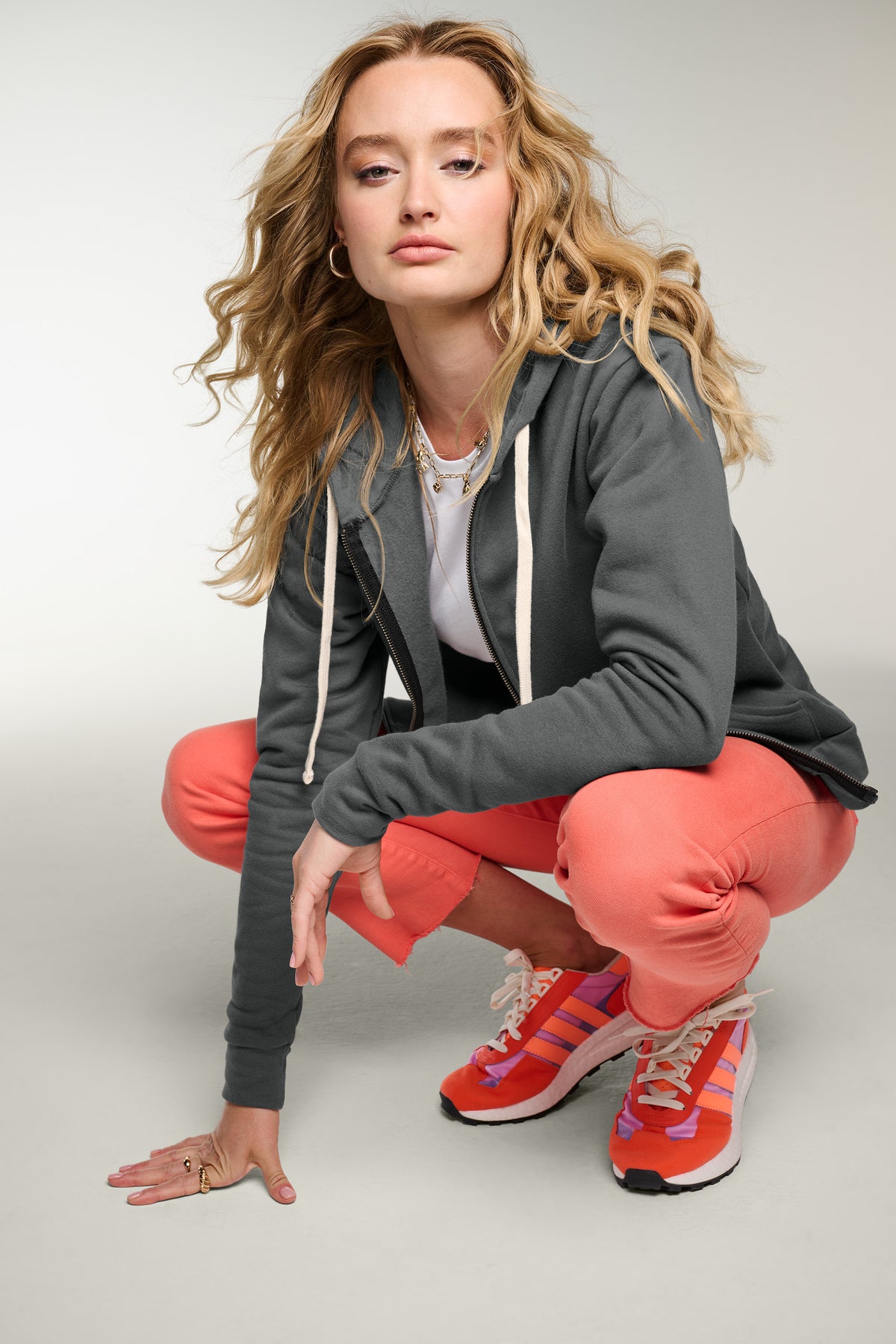A woman crouching in a studio setting, wearing a dark gray zip-up hoodie, coral pink joggers, a white top, and red and pink athletic sneakers. Next Level Apparel® Adult Santa Barbara Zip Hoodie, Embroidered
