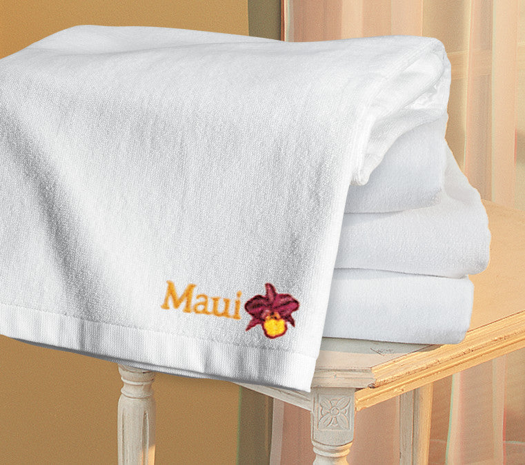 Stack of white towels with 'Maui' embroidery on a wooden chair. Port Authority® Cabana Beach Towel, Embroidered by Helmsman Stitch Co.