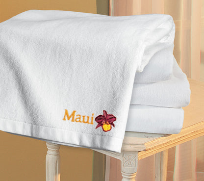 Stack of white towels with 'Maui' embroidery on a wooden chair. Port Authority® Cabana Beach Towel, Embroidered by Helmsman Stitch Co.