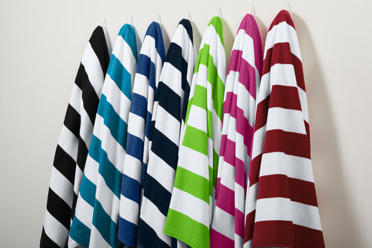 Set of colorful striped beach towels hanging against a plain background. Port Authority® Cabana Stripe Beach Towel, Embroidered by Helmsman Stitch Co.