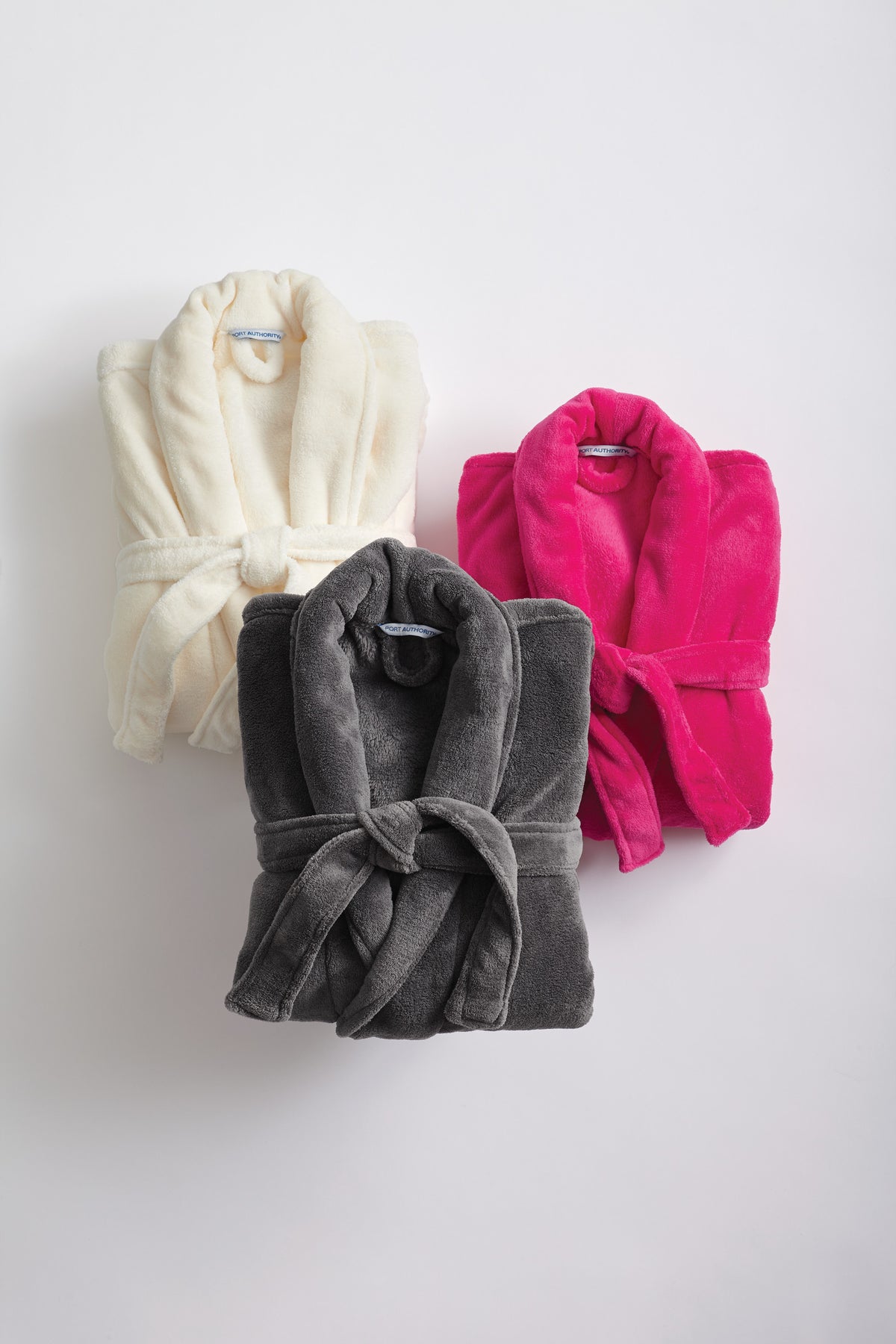 Three plush bathrobes arranged on a light gray background: one cream, one dark gray, and one bright pink. The gray robe is in front and centered. Port Authority® Plush Microfleece Shawl Collar Robe, Embroidered