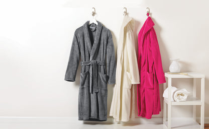 Three plush bathrobes hang on white hangers against a white wall: a gray robe on the left, a cream robe in the center, and a bright pink robe on the right. A small white side table with a folded towel and white vase sits to the right. Port Authority® Plush Microfleece Shawl Collar Robe, Embroidered