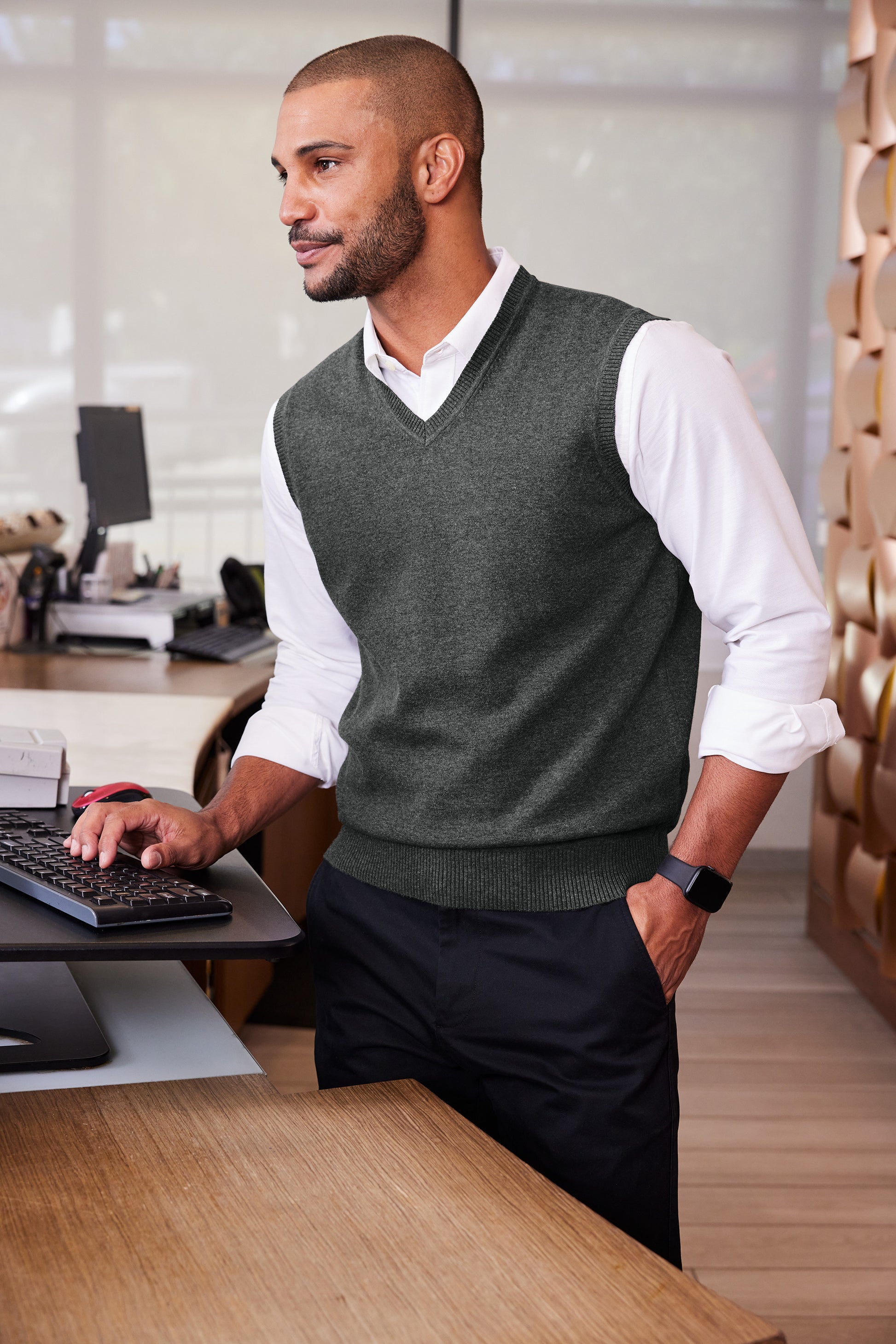 A man in a modern office setting wearing a dark gray v-neck knit sweater vest over a white collared shirt and dark trousers, standing next to a desk. Port Authority® Easy Care Sweater Vest, Embroidered
