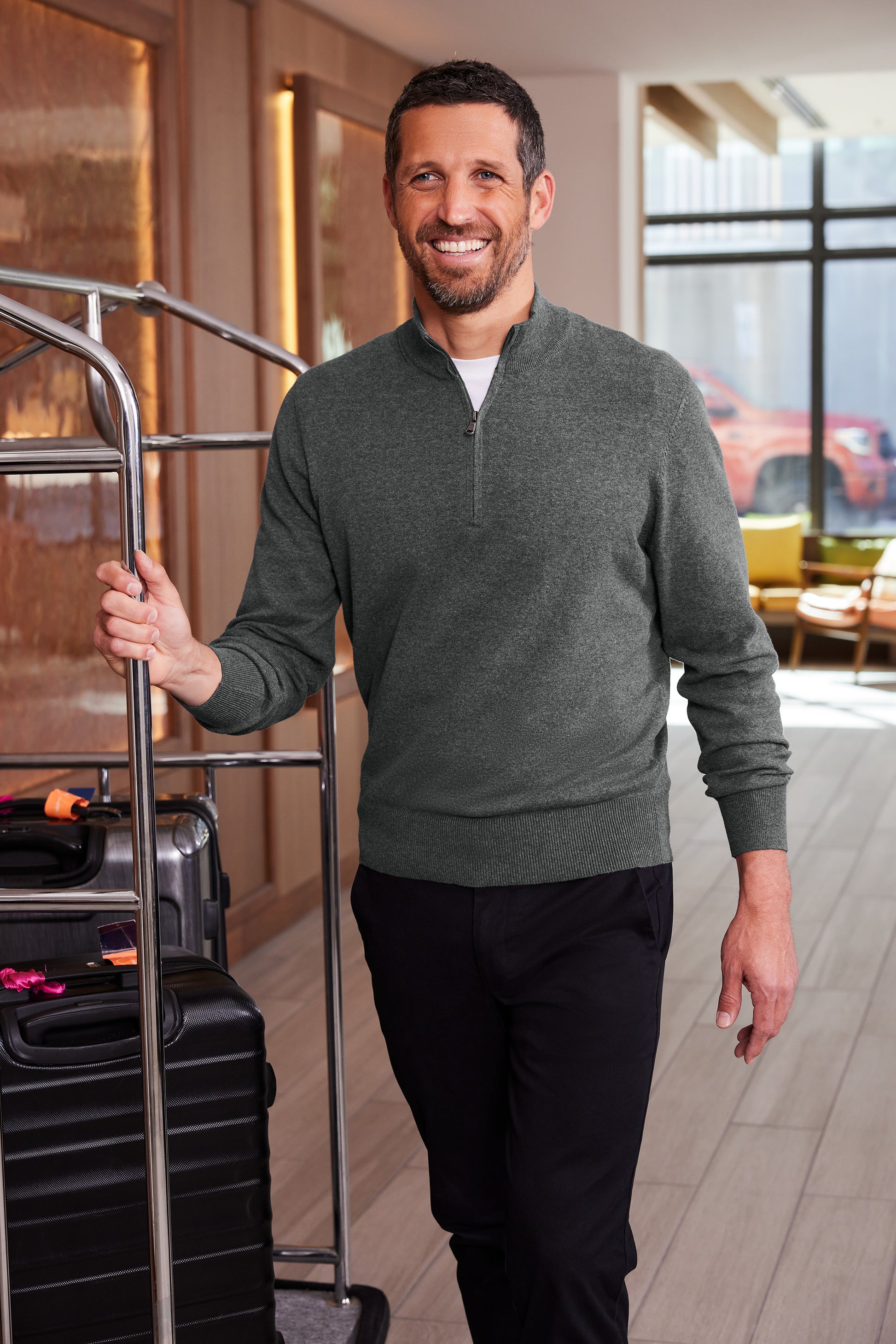 A smiling man wearing a dark green quarter-zip pullover sweater and black pants stands in a hotel lobby next to a luggage cart with suitcases. Port Authority® Easy Care 1/4-Zip Sweater, Embroidered