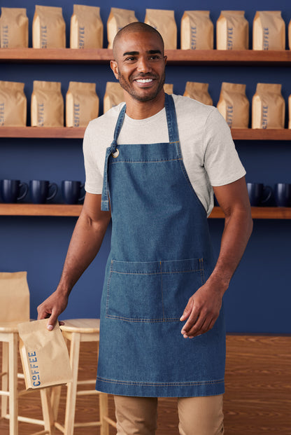 Man wearing a blue apron in a coffee shop setting with shelves of coffee bags in the background. Port Authority® Signature Denim Apron, Embroidered
